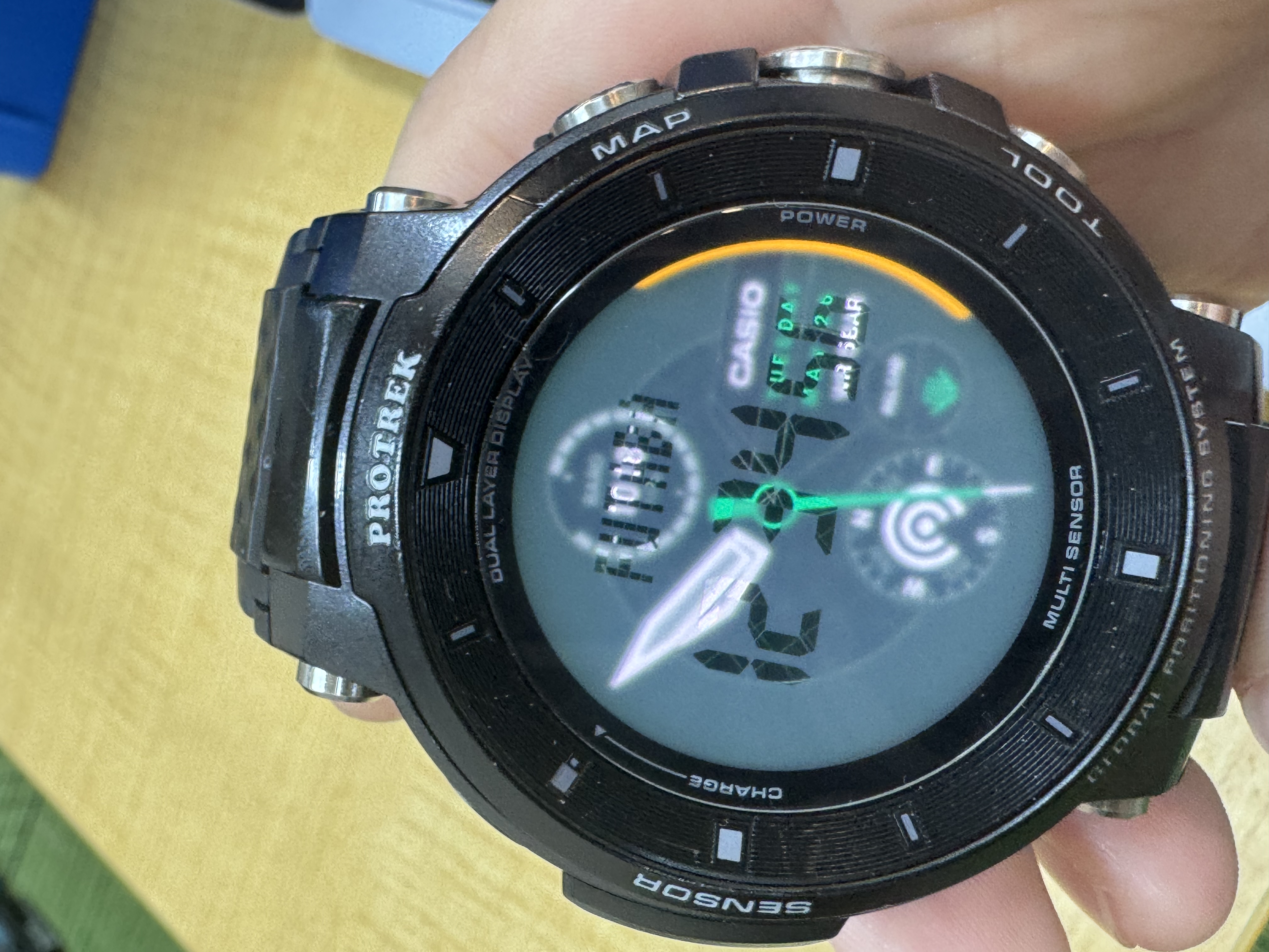 Casio WSD-F30 で dual-layer 表示が成立している実機写真。背景に Wear OS の watchface (アナログ針 + デジタル時計 12:34、TUESDAY MAY 26、コンパス、気圧計) が出ているところに、サブLCDの「FUTABA」(上部マトリクス) と「12 34 56」(中央 7-seg) が同時に重なって見えている